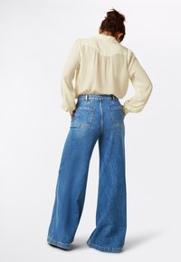 Blusa color crema con yoke e una consistenza morbida, abbinata a jeans in denim blu a gamba larga con tasche posteriori e un lavaggio sbiadito.