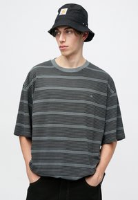 Camiseta de rayas grises con un corte relajado, mangas cortas y cuello acanalado, combinada con un sombrero de cubo negro que presenta un parche con logo.