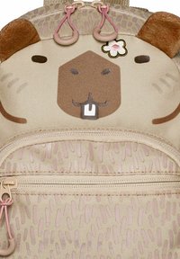 Mochila beige con diseño de cara de capibara, textura suave, orejas redondas y detalles florales. Incluye un bolsillo delantero con cremallera.