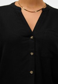 Chemise noire à boutons en tissu tissé avec une poche, dotée d'un décolleté en V et de boutons marron clair sur le devant.