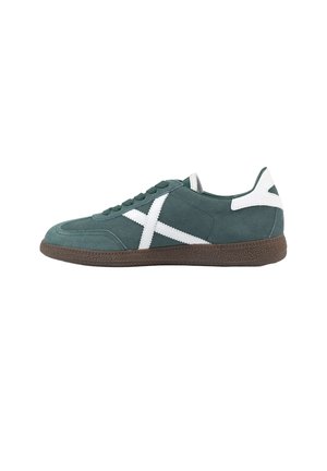Zapatilla verde de ante con rayas blancas cruzadas, detalle blanco en el talón, suela de goma marrón y cordones, mostrada desde el lado izquierdo sobre un fondo blanco.
