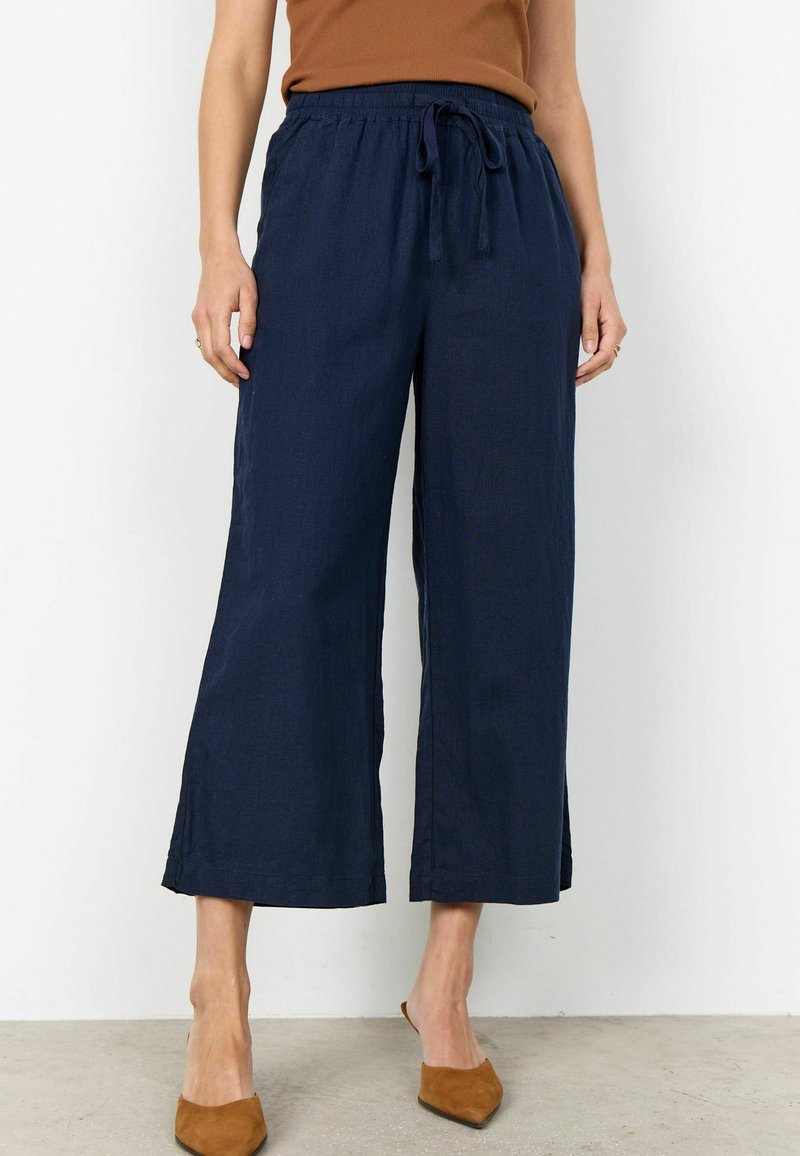 Femme portant un pantalon large court bleu marine avec taille élastique et cordon de serrage, assorti à des talons bruns à bouts pointus et un haut rouille.