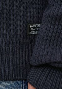 Navy ribbed sweater featuring a textured knit design, label detailing, and a snug fit at the cuffs, made from soft fabric.