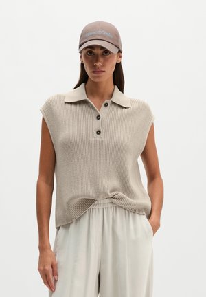Vrouw draagt een beige gebreide mouwloze polo, lichte losse broek en een taupekleurige baseballpet met tekst, staand met één hand in de zak.
