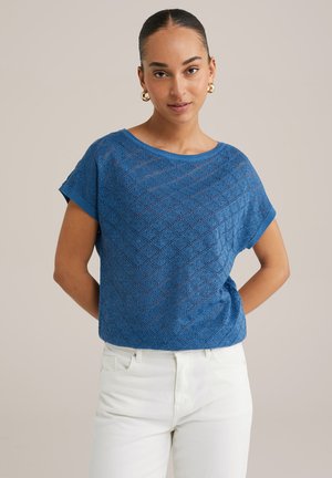 Blauw gebreid T-shirt met korte mouwen, voorzien van een ruitpatroon, ronde halslijn en een ontspannen pasvorm, gedragen met witte broek op een neutrale achtergrond.