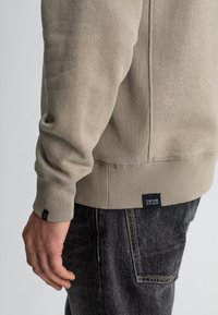 Beige katoenen sweatshirt met ribgebreide manchetten en zoom, voorzien van een zijnaad en een label op de taille. Gepaard met donkere denimjeans.