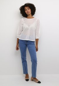 Witte texturen blouse met 3/4 mouwen, met een geometrisch patroon. Gecombineerd met lichtblauwe jeans en beige blokhak-schoenen met zwarte punten.