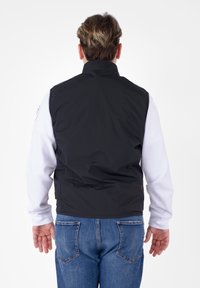 Gilet nero senza maniche sopra una camicia bianca a maniche lunghe, con colletto alto e texture liscia, abbinato a jeans in denim blu.