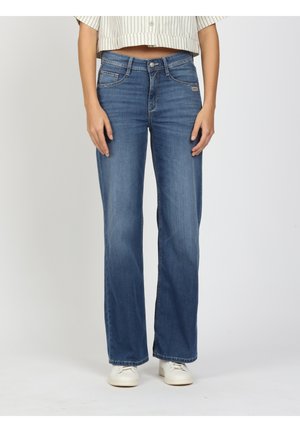 Frau trägt Jeans mit geradem Bein in mittlerer Waschung, mit Vordertaschen und weißen Turnschuhen, steht vor hellgrauem Hintergrund.