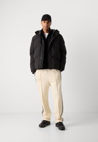 Zwarte pufferjas met een rits, grijze binnenkant, zwarte hoodie eronder, crèmekleurige broek met zijdelingse strepen en zwarte sneakers.
