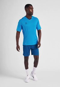 Herre træningsoutfit med en blå kortærmet skjorte og matchende shorts. Skjorten har rund hals og logodetalje; stoffet virker glat.