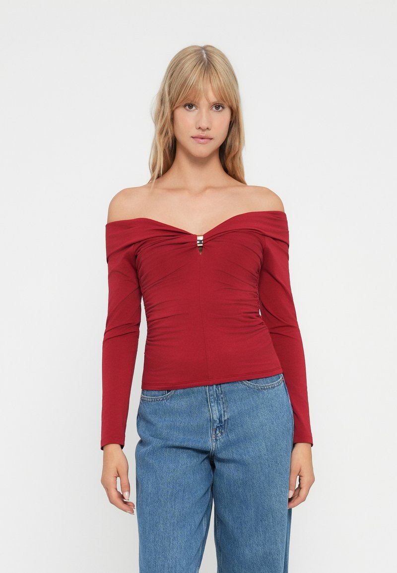 Top rojo sin mangas, hecho de tela elástica, con mangas largas, un diseño fruncido y un detalle central. Combinado con jeans azules.
