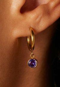 Goldene Creolen mit einem hängenden, runden lila Edelstein. Der Edelstein ist facettiert und fängt das Licht ein, und das Ohrläppchen ist im Hintergrund sichtbar.