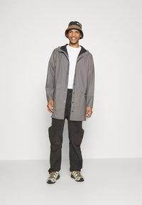Grå vattentät jacka med huva, knappstängning och två framfickor. Bärs över en vit skjorta och i kombination med mörka cargo-byxor och sneakers.