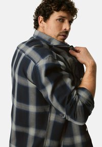 Niebiesko-szara koszula w kratę z zapiętym kołnierzykiem na guziki i podwiniętymi rękawami, wykonana z teksturowanej tkaniny z widocznymi szwami.