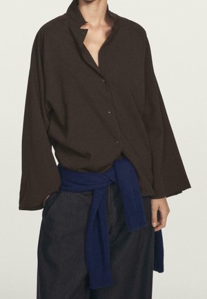 Personne portant une chemise boutonnée ample marron foncé à manches larges, un pantalon sombre et un pull bleu noué autour de la taille, debout devant un fond uni.