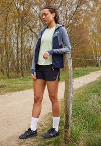 Žena v atletickém oblečení se opírá o dřevěný sloupek plotu podél pěšiny v lesnaté oblasti během podzimu.