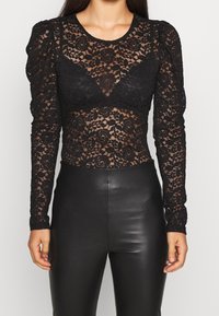 Haut noir à manches longues en dentelle avec motifs floraux, design transparent et épaules bouffantes, associé à un pantalon en cuir noir taille haute.