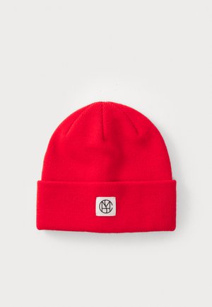Red knit beanie with a folded cuff and a square logo patch featuring black letters on a white background. Textured fabric.