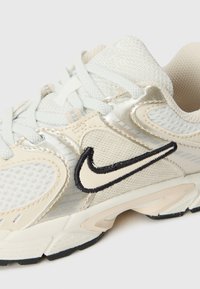 Nike løpesko med et lett beige nettingoverdel, reflekterende sølvdetaljer, svart brodert logo og teksturert gummisåle.