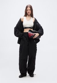 Νεαρή γυναίκα με μαύρο παντελόνι cargo, λευκό κοντό τοπ, φαρδιά μαύρη ζακέτα, κρατώντας μαύρο και κόκκινο clutch σε απλό φόντο.