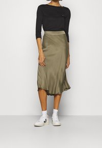 Una donna è in piedi in una gonna verde oliva elegante e una maglietta nera, abbinata a sneaker bianche, evidenziando un outfit casual ma alla moda.