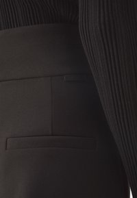 Gros plan sur un pantalon noir avec une poche arrière et une manche de pull noir côtelée visible sur le côté droit.