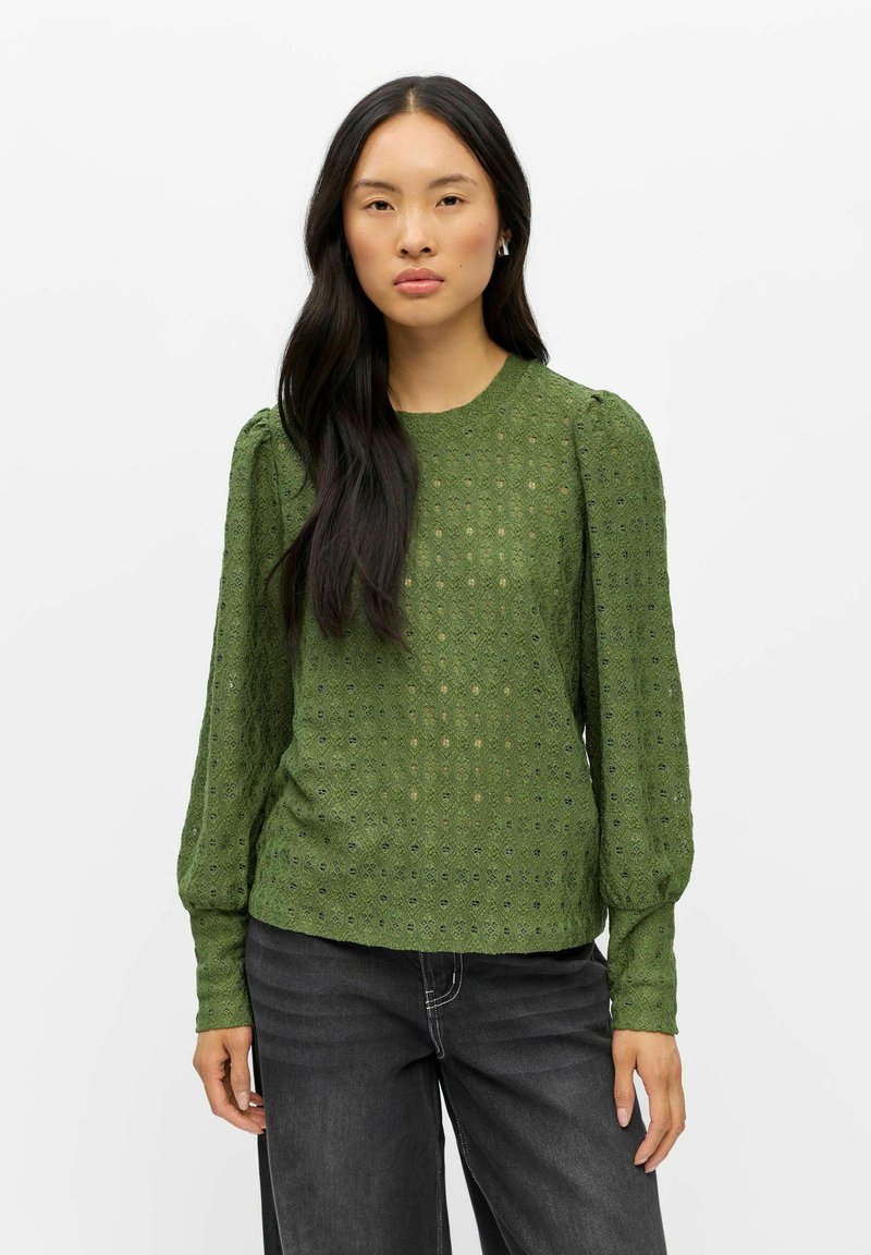 Femme aux longs cheveux noirs portant un chemisier vert texturé à manches longues et un jean gris foncé, debout devant un fond blanc uni.