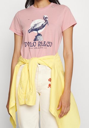 Osoba nosząca różowy t-shirt z grafiką pelikana i napisem, żółtą koszulę zawiązaną w pasie oraz kremowe spodnie z małym czerwonym haftem.