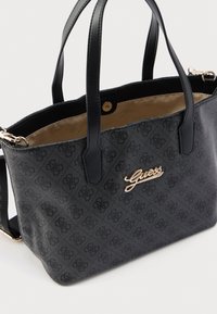Čierna taška typu tote s reliéfnym vzorom loga, zlatými kovovými doplnkami, dvoma pevnými rukoväťami a béžovou vnútornou podšívkou so spoľahlivým zapínaním na cvok.