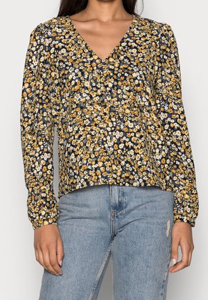 Blus med blommönster i marinblått, med gula och vita blommor. Den har V-ringning och långa ärmar med rynkade muddar.