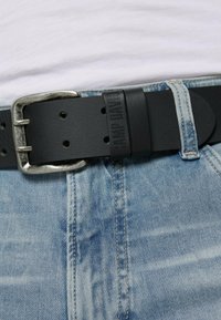 Schwarzer Ledergürtel mit silberner Metallschnalle, ausgestattet mit mehreren Löchern zur Anpassung. Getragen mit hellblauen Jeans.