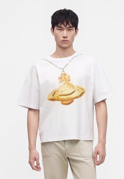 Νέος άνδρας φοράει λευκό oversized t-shirt με μεγάλο χρυσό γραφικό πλανήτη και σταυρό και μπεζ παντελόνια σε απλό φόντο.