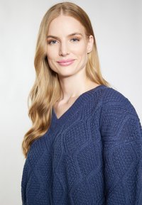 Granatowy sweter z dzianiny z dekoltem w serek, teksturowanym heksagonalnym wzorem oraz prążkowanymi ściągaczami, z długimi włosami częściowo widocznymi w kadrze.