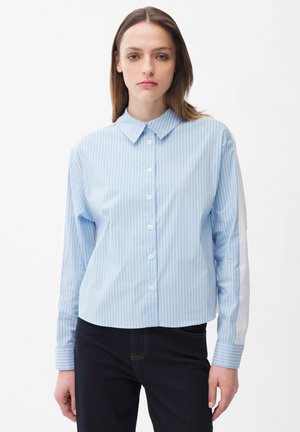 Jeune femme portant une chemise boutonnée bleu clair à rayures verticales avec un détail de rayures blanches sur les manches et un pantalon foncé, faisant face à l'avant.