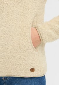 Beige fleece pullover with soft, textured fabric, side pocket, and contrasting trim. Features a small brown logo tag near the hem.