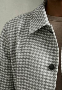 Abrigo de patrón de espiga gris con una textura suave, que presenta un cuello puntiagudo y detalles de botones negros en el frente.