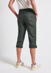 Groene cropped broeken met een elastische tailleband, opgerolde mouwen en achterzakken. Zachte textuur en duurzaam materiaal. Voeten in witte sneakers.