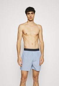 Boxer (slip) a righe blu e bianche realizzato in tessuto leggero, con una cintura elastica nera e una vestibilità ampia.
