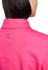 Leuchtend pinke Sportjacke mit weicher Textur, ausgestattet mit einem Kragen und einem sichtbaren Etikett, auf dem "Marc Cain Sports" steht. Die Rückansicht hebt die Designdetails hervor.