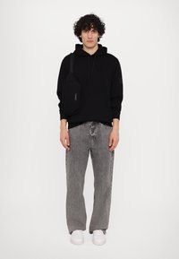 Felpa nera in cotone, jeans in denim grigi, e sneakers bianche. Il modello porta una borsa a tracolla nera con un design minimale e un marchio discreto.