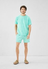 Camiseta de color teal con una textura suave combinada con pantalones cortos de teal con un diseño geométrico, complementados con sandalias marrones.