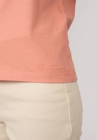 T-shirt en coton couleur pêche à manches courtes, avec un ourlet incurvé et un logo brodé en rose clair sur le côté.