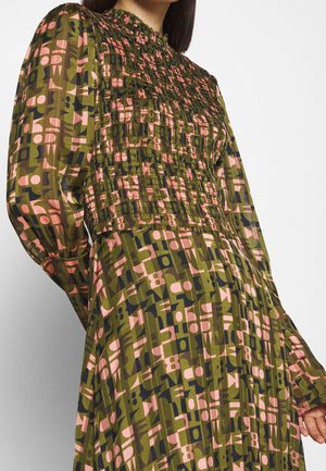 Frau trägt ein olivgrünes, langärmliges Kleid mit rosa und marineblauen geometrischen Mustern sowie einem gerafften, hochgeschlossenen Ausschnitt und Oberteil.