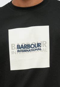 Fekete pamut póló, fehér téglalap alakú grafikával, rajta kék "BARBOUR" és szürke "INTERNATIONAL" felirat, texturált mintával.