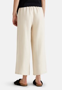 Pantalones beige de pierna ancha con cintura elástica, hechos de un tejido ligero, presentan una textura suave y un corte recto. Se llevan con zapatillas negras.