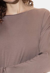 Vrouw in een mauve langemouwtop met een bootnek, die een deel van haar krullend haar en nek toont.