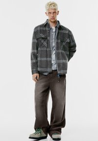 Šedá károvaná flanelová bunda s dvoma náprsními vreckami cez károvanú košeľu, skombinovaná s hnedými kordovou nohavicami a textúrovanou obuvou so zelenými akcentmi.