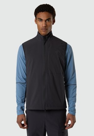 Uomo che indossa un gilet nero senza maniche con cerniera sopra una camicia blu a maniche lunghe, in piedi davanti a uno sfondo grigio chiaro uniforme.