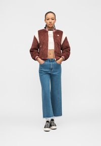 Νεαρή γυναίκα στέκεται με τα χέρια στις τσέπες, φοράει μπουφάν φλις σε μπορντό και λευκό, λευκό κοντό τοπ, φαρδιά μπλε τζιν παντελόνια και μαύρα αθλητικά παπούτσια.